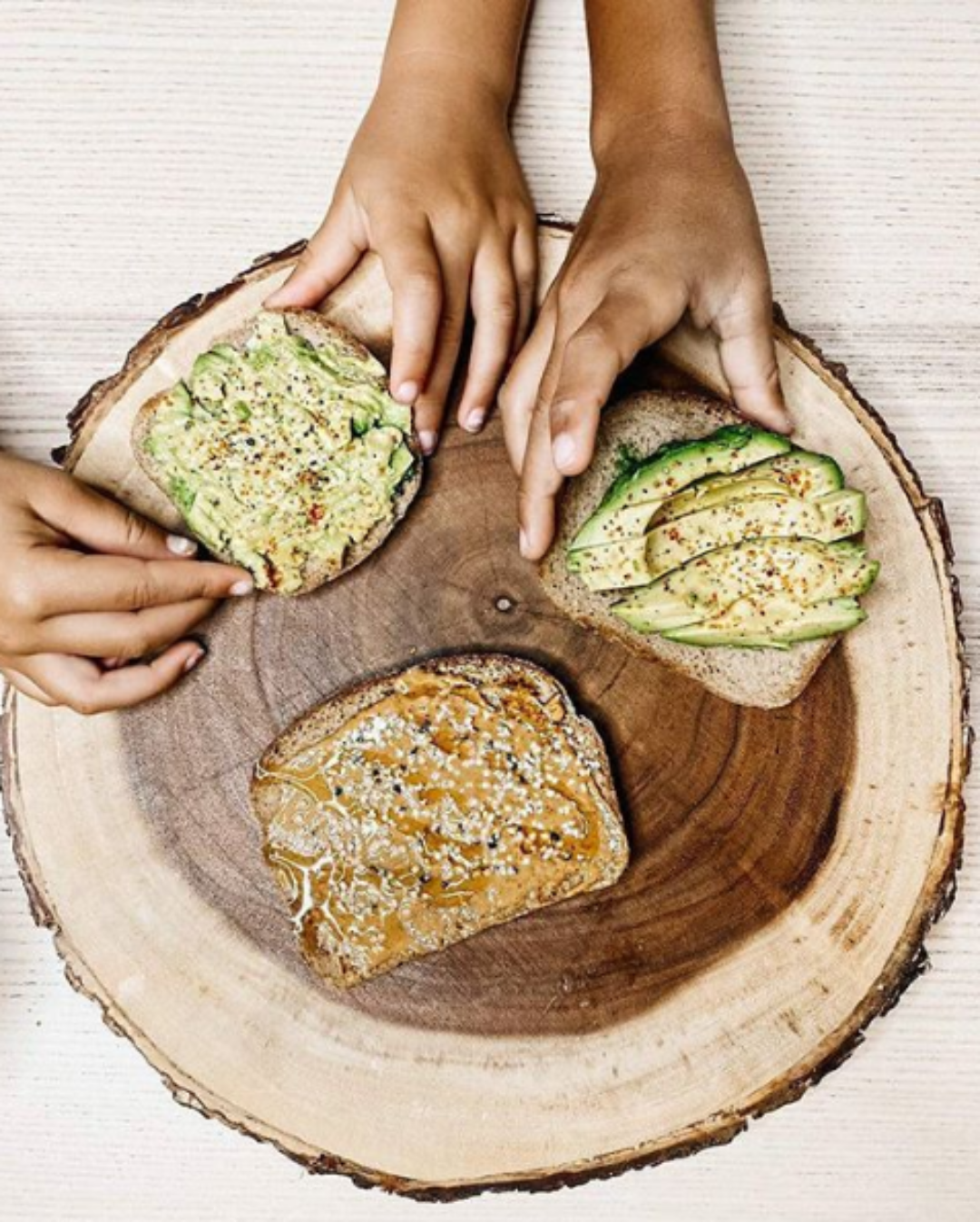 Two slices of avocado toast on a wooden board with two hands reaching for them.