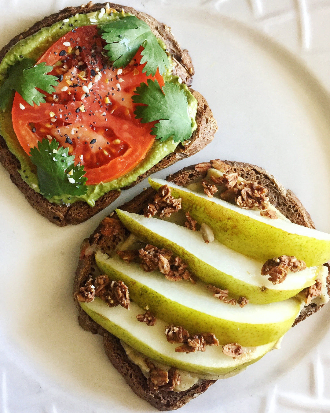 Two slices of bread with avocado, tomato, and cilantro on one, and pear slices and granola on the other, on a white plate.