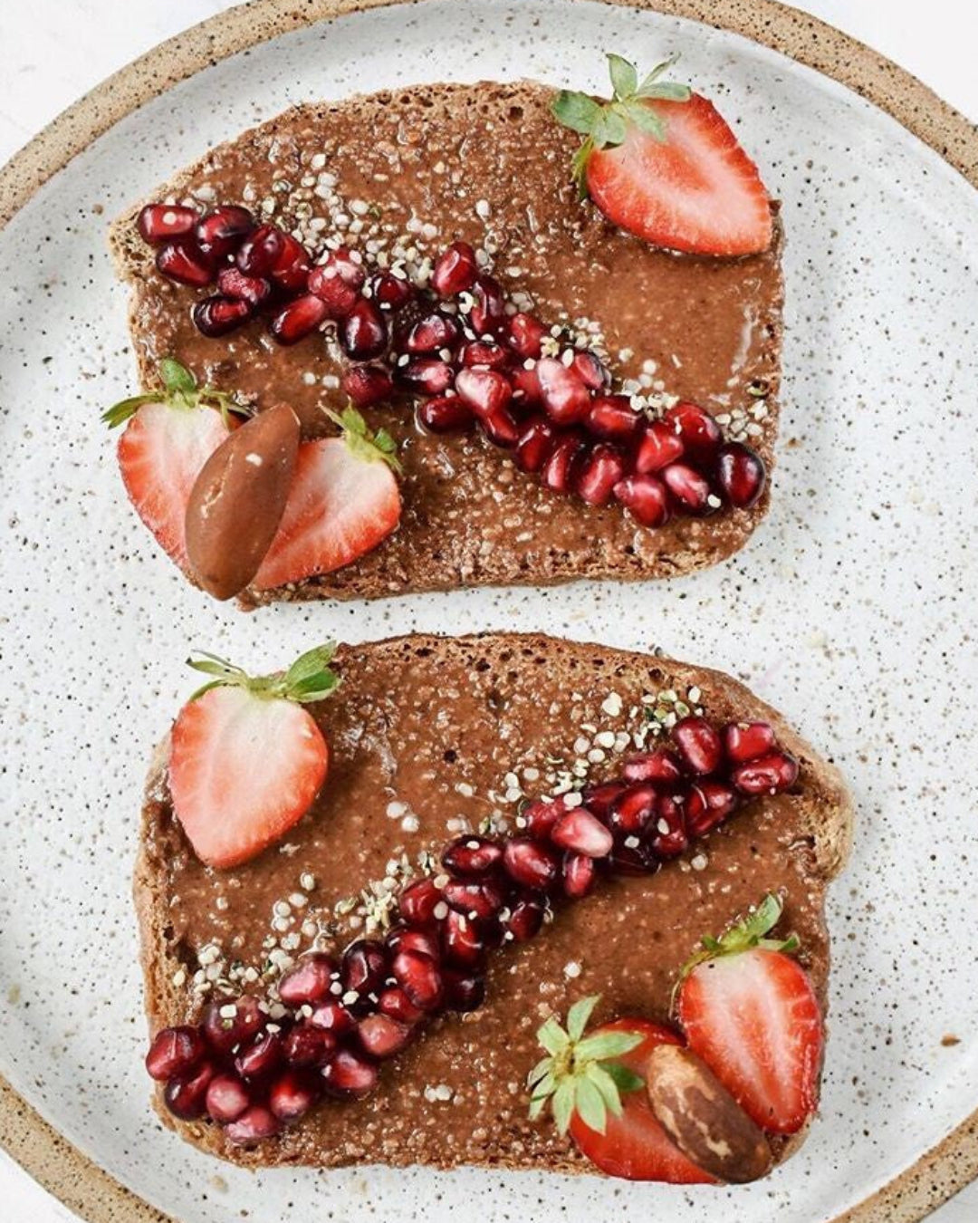 Two pieces of toast with nutella, strawberries, and pomegranate seeds on a speckled plate.
