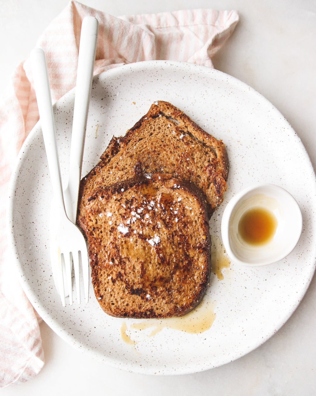 Two slices of French toast on a white plate with syrup and a fork, on a light background.