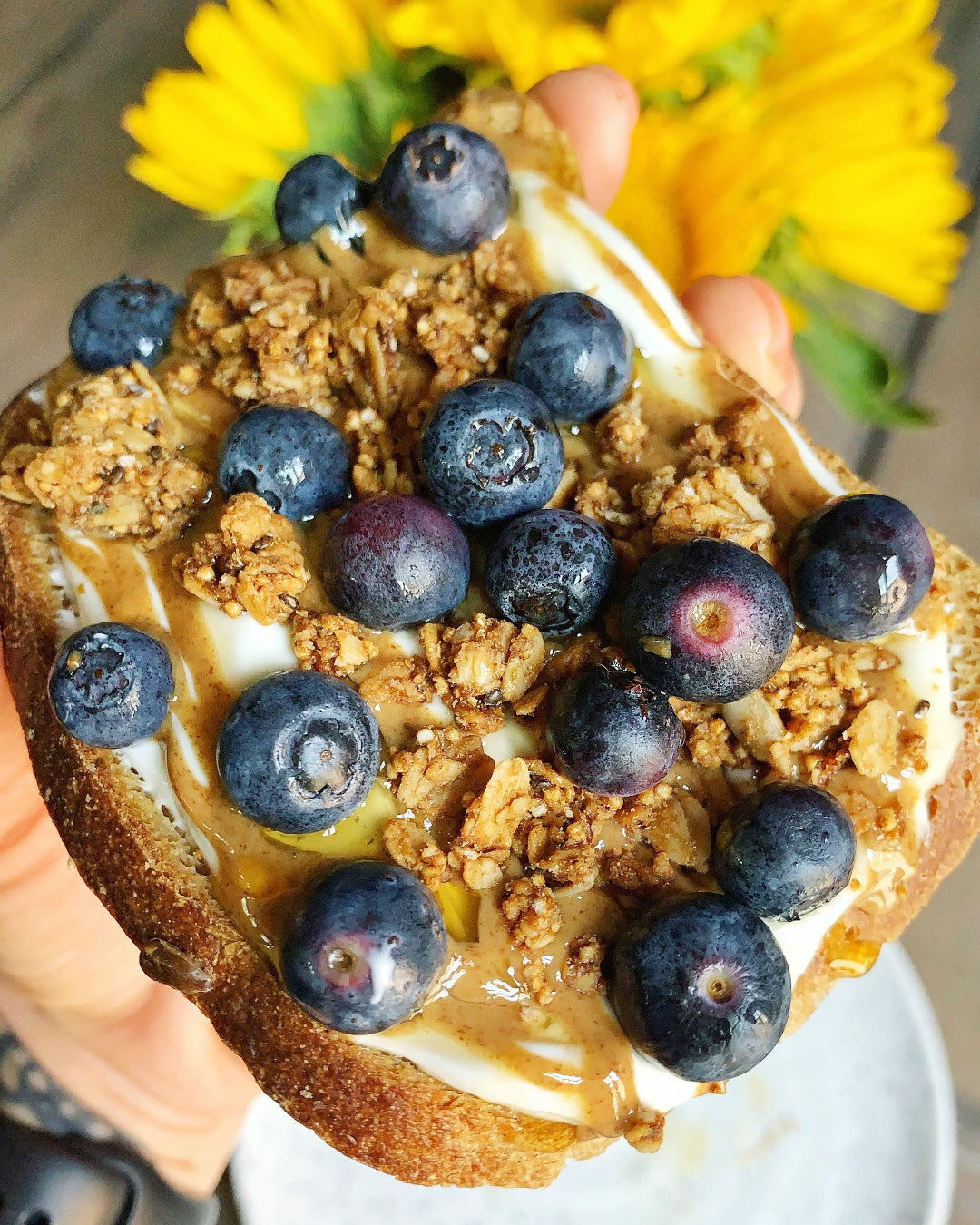 Hand holding a toast with blueberries, granola, and syrup against a blurred background of yellow flowers.